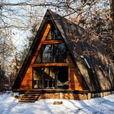 An Aframe cabin in the woods with snow on the ground and firewood under the front porch