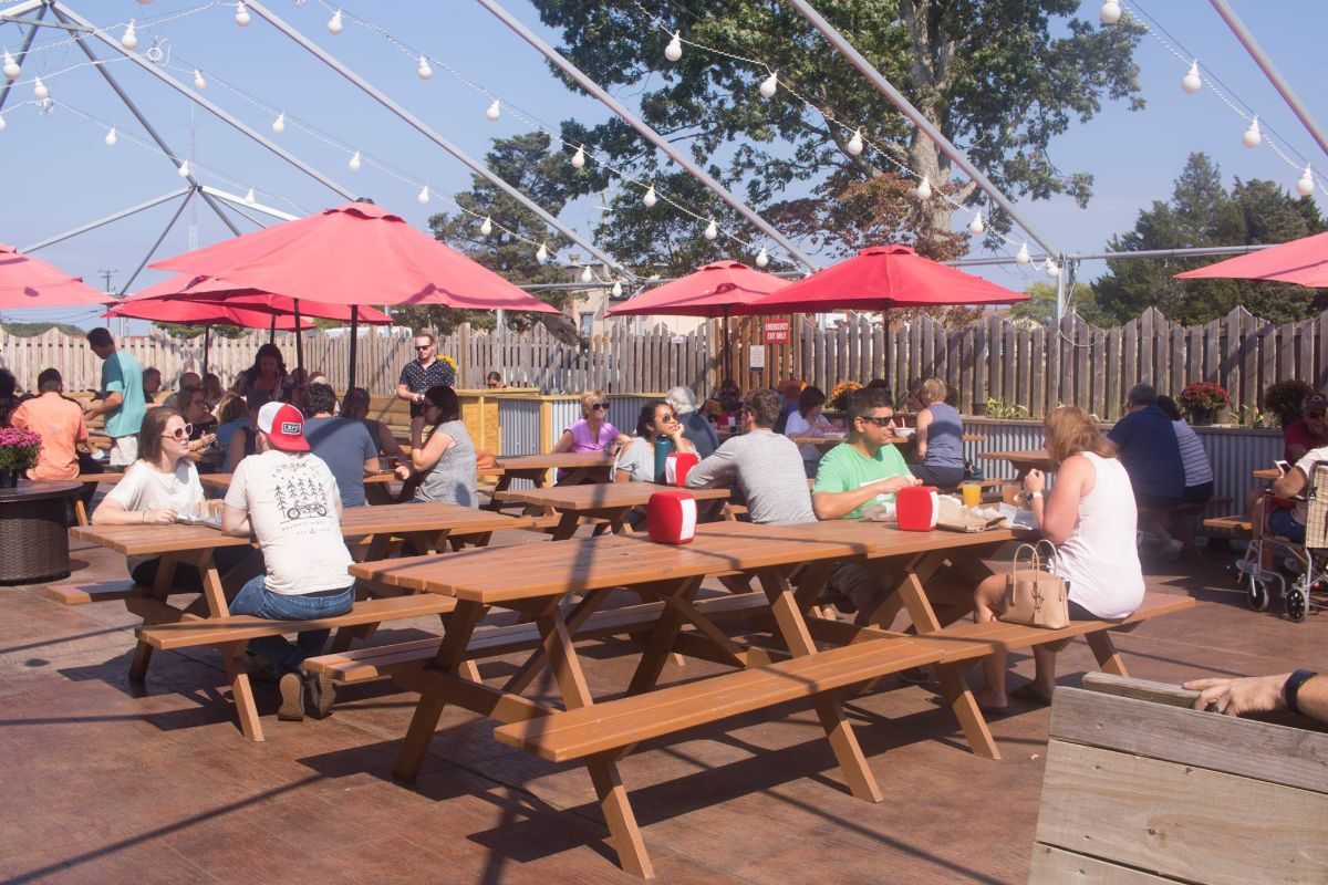 Beer drinkers sitting at many picnic tables at Cape May Brewing Cos outdoor seating area at their tasting room with umbrellas and string lights