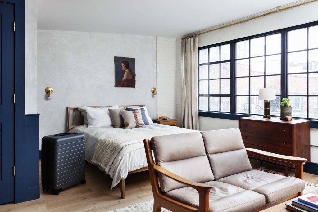 The top floor Ben Suite at Lokal Hotel Old City with queen bed and vintage leather love seat with mid century modern dresser next to huge industrial windows flooding room with light