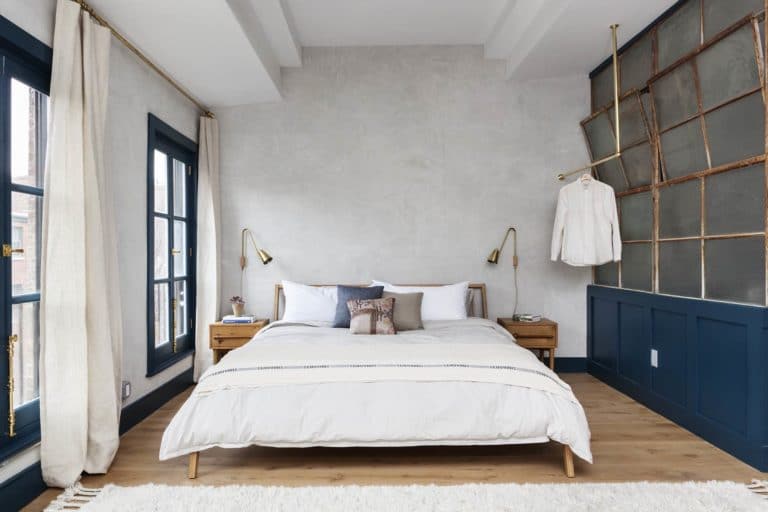 King bed flooded with light from floor to ceiling windows and brass task lighting on either side with clothes hanging from an exposed brass rod next to a reclaimed factory window divider