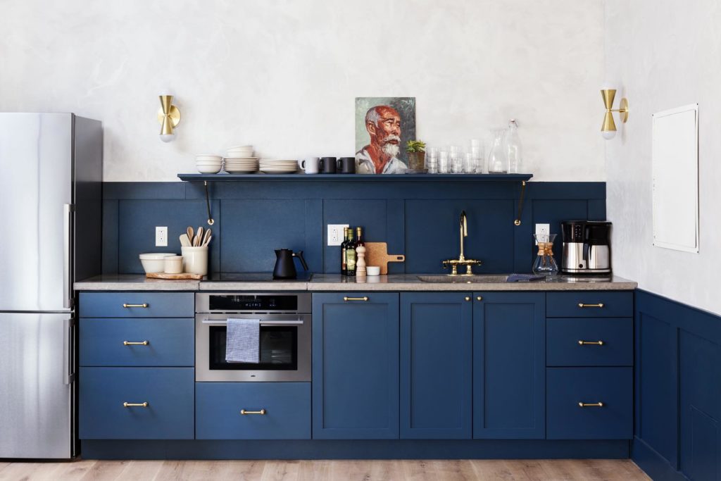 Bright blue kitchen with concrete countertops and floating shelf with brass hardware and sconces and an oil painting of man on shelf