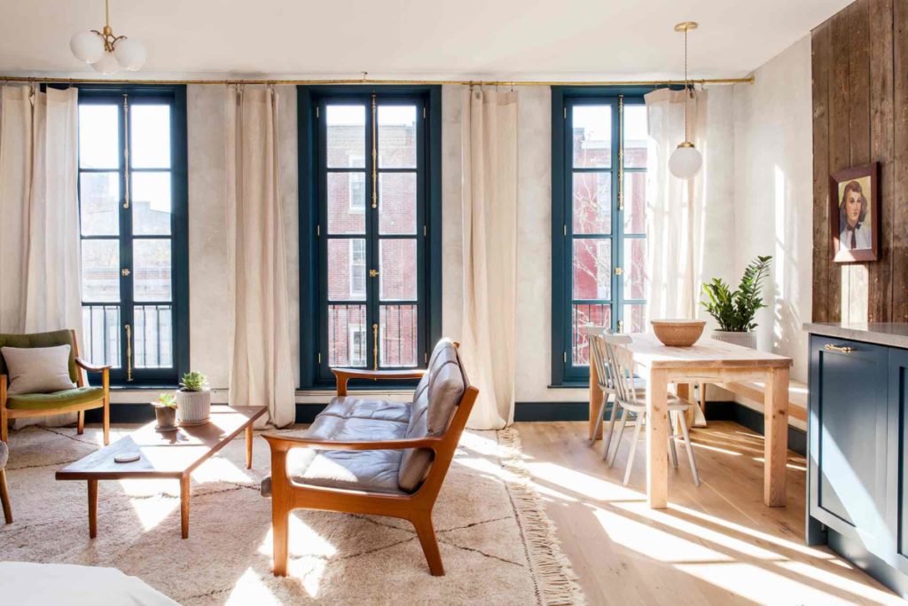 Three floor to ceiling historic windows pour light into this Lokal Hotel apartment suite in Old City with Mid Century Modern furniture in the living room and custom made wood dining table at the end of the kitchen with wide plank white oak floors and a vintage rug