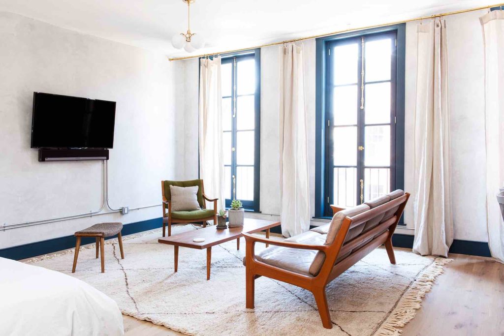 The living room of the Tina suite in Lokal Hotel Old City is flooded with natural light by historic operable floor to ceiling windows and contains vintage leather furniture on a plush area rug with smart tv and sonos sound
