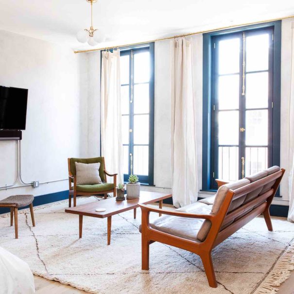 The living room of the Tina suite in Lokal Hotel Old City is flooded with natural light by historic operable floor to ceiling windows and contains vintage leather furniture on a plush area rug with smart tv and sonos sound