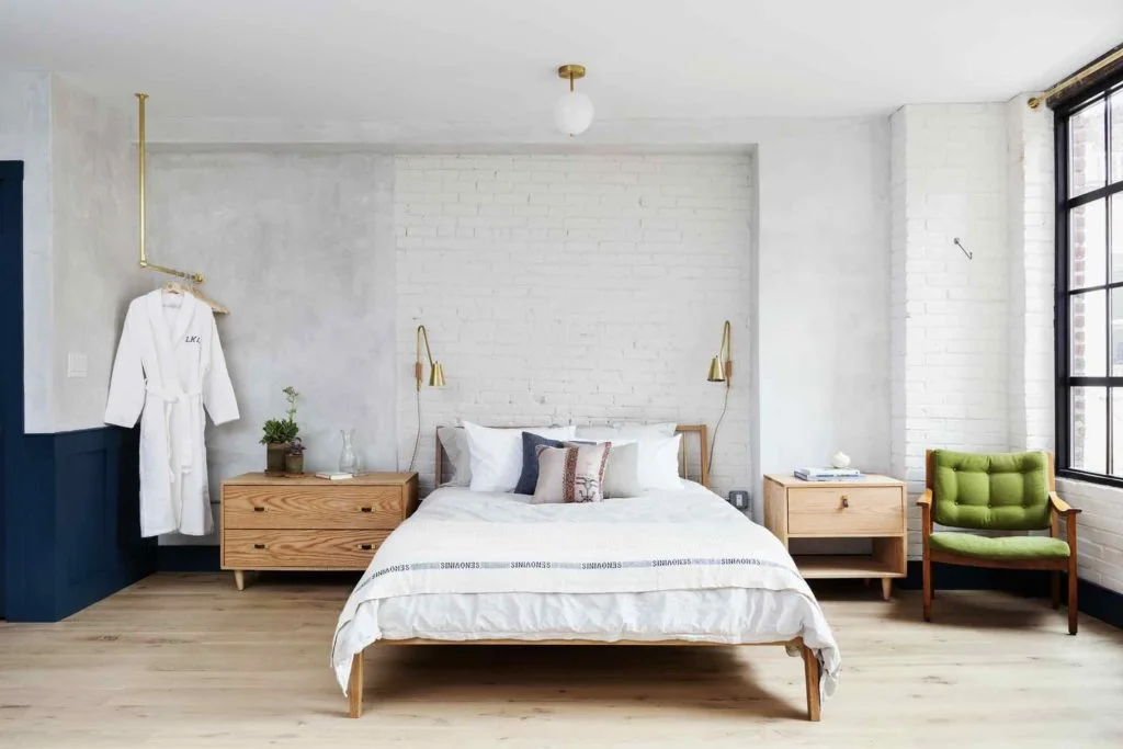 A neatly made bed with white linens and pillows in a modern bedroom with a green chair and white robe.