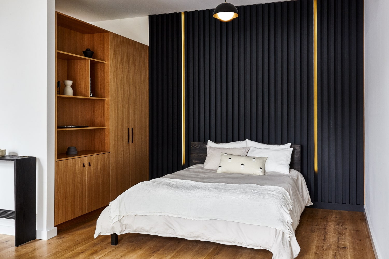 A neatly made bed with white linens and pillows against a black wall with gold accents, with a wooden cabinet and shelves to the left.
