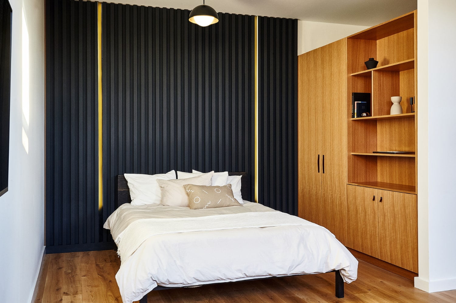 A neatly made bed with white linens and pillows against a black wall with gold accents, with a wooden cabinet and shelves to the right.