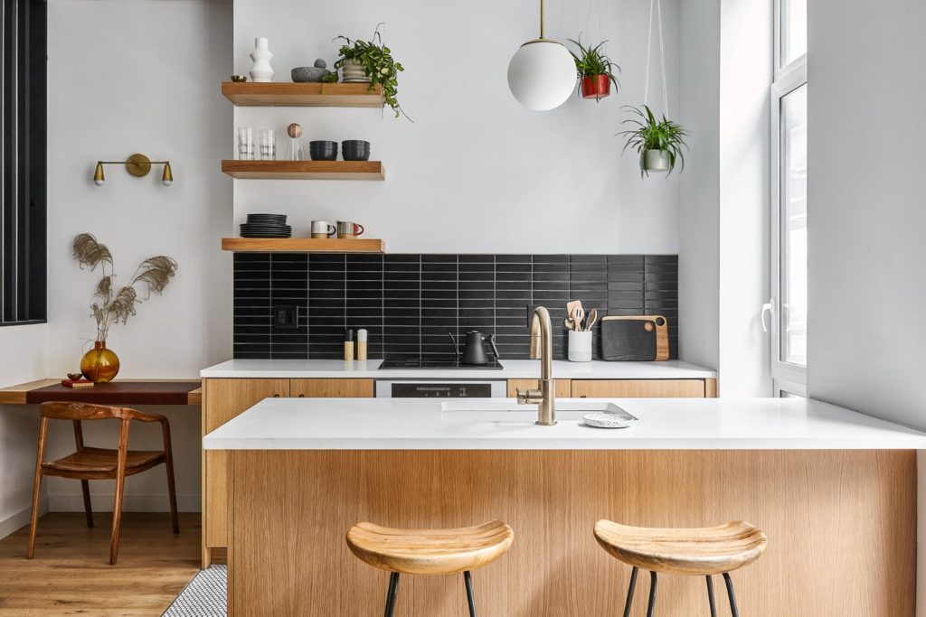 white oak kitchen with island and white quartz countertops floating shelves and brass light fixture with hanging plants in the corner and two stools behind the island and ADA sitting desk to the left
