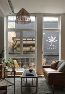 View of the Garden Jawn living room at Lokal Hotel Fishtown looking over the garden through the windows in the back of the property with seating, trellis and logo painted on the back wall