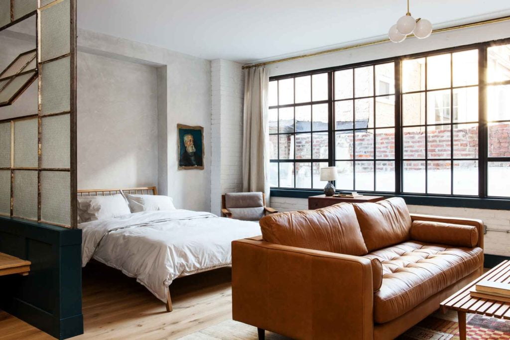 Light pouring into huge warehouse windows into this apartment hotel room by Lokal Hotel in Old City with leather couch in the foreground and queen bed with parachute home linens in background and oil painting on plaster walls