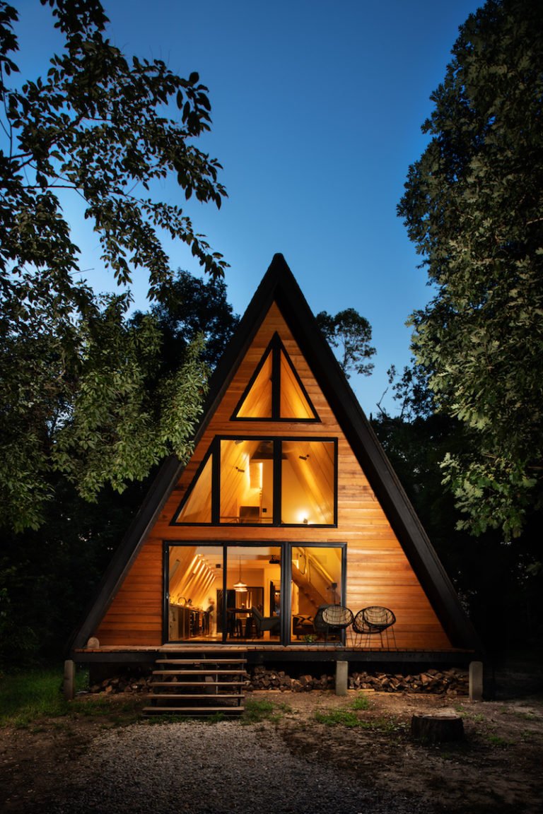 An Aframe cabin in the woods at night with warm lights glowing through front facade of glass