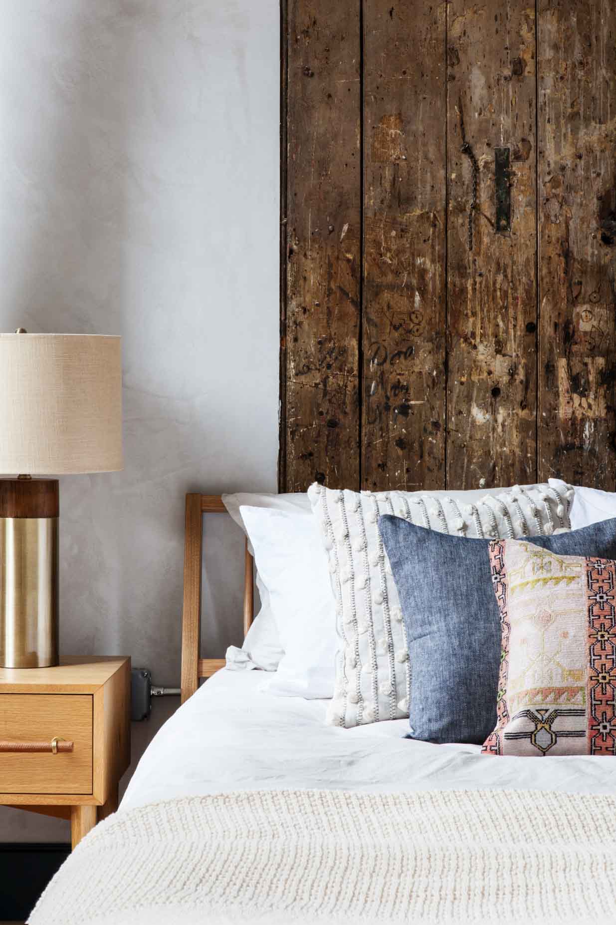 A bed with white linens and several pillows is positioned against a wall with wooden paneling, accompanied by a wooden nightstand and lamp.