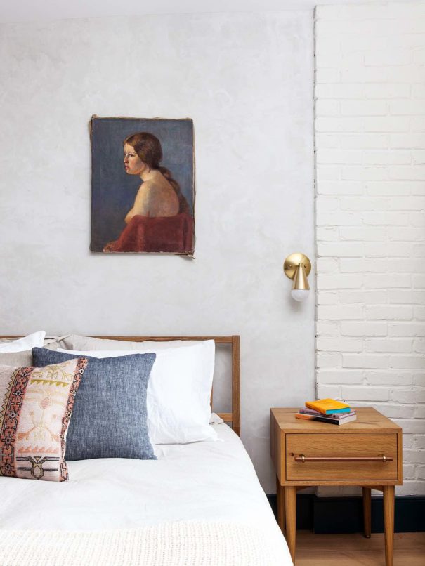 hotel bed with colorful throw pillows a mid century side table and an oil painting hanging on white painted exposed brick