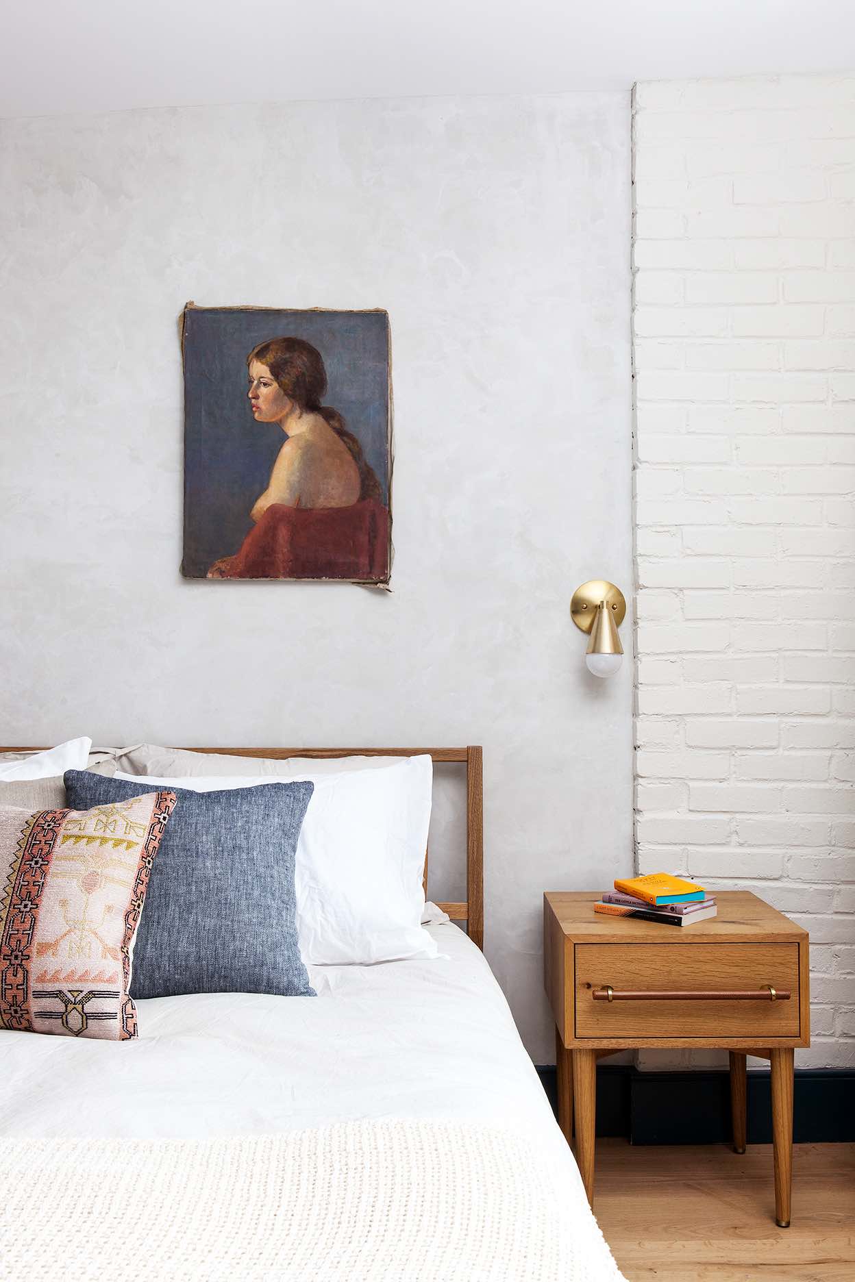 A neatly made bed with white linens and colorful pillows, a wooden nightstand with books, and a painting of a woman on the wall in a white room with a brick accent wall.