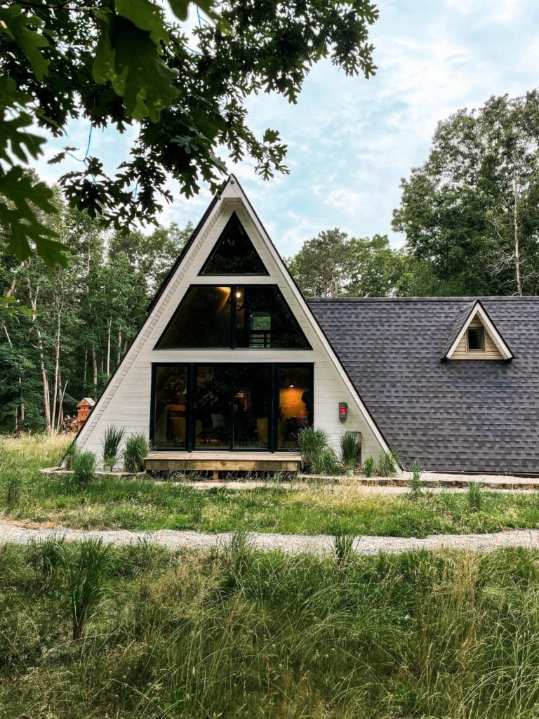 Aframe Triangle House with white siding and large glass wall with path winding through landscape in front of it